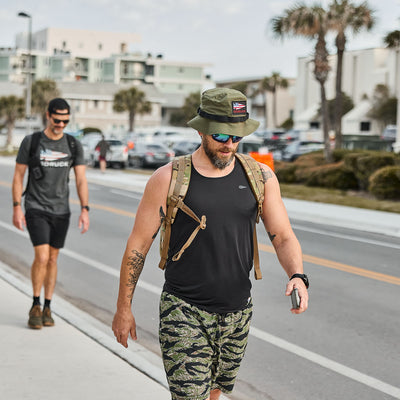 A man in a black tank top and camo shorts, wearing a ToughDry Tactical Boonie Hat, walks outdoors holding a phone. Another person follows him.