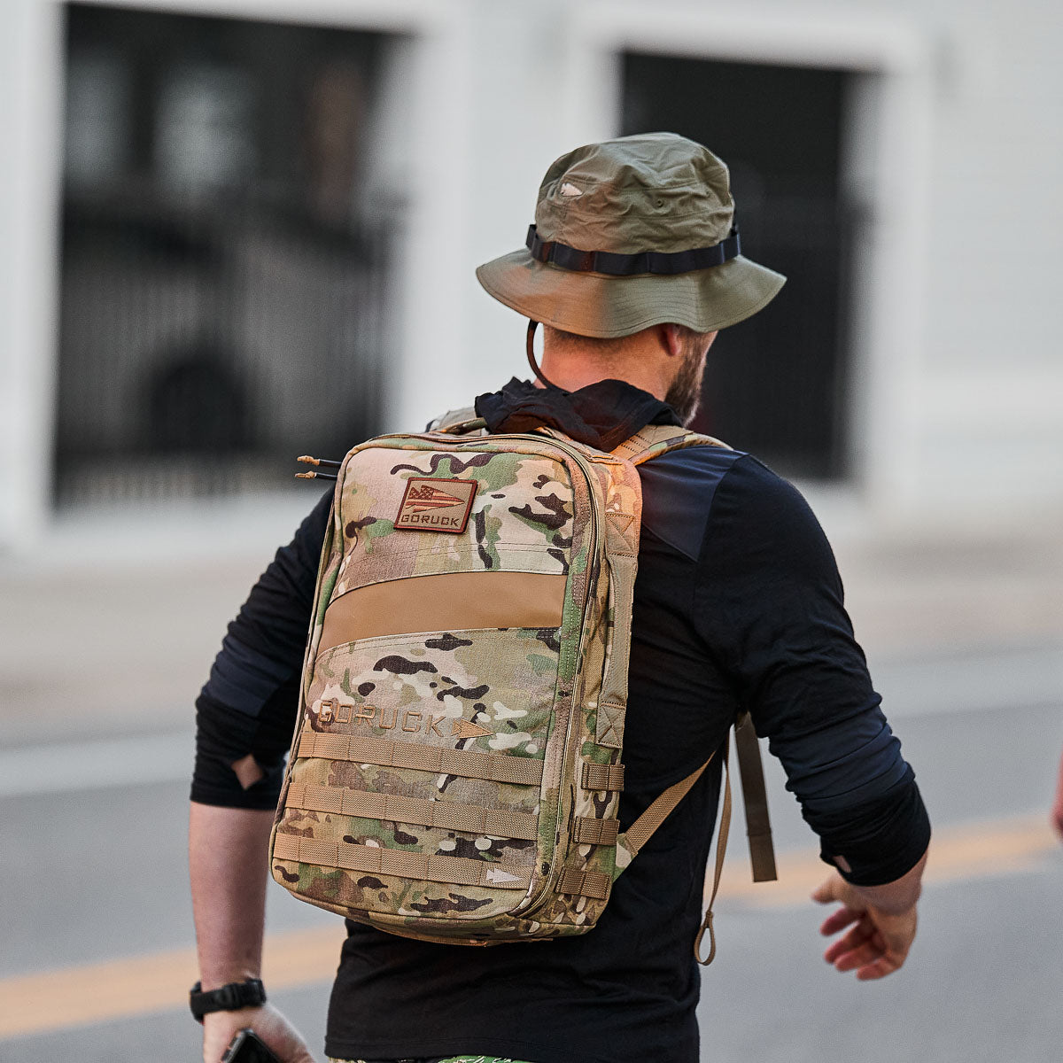 A person sporting a military-style camouflage backpack and a Boonie Hat - Tactical - ToughDry strolls down the street.