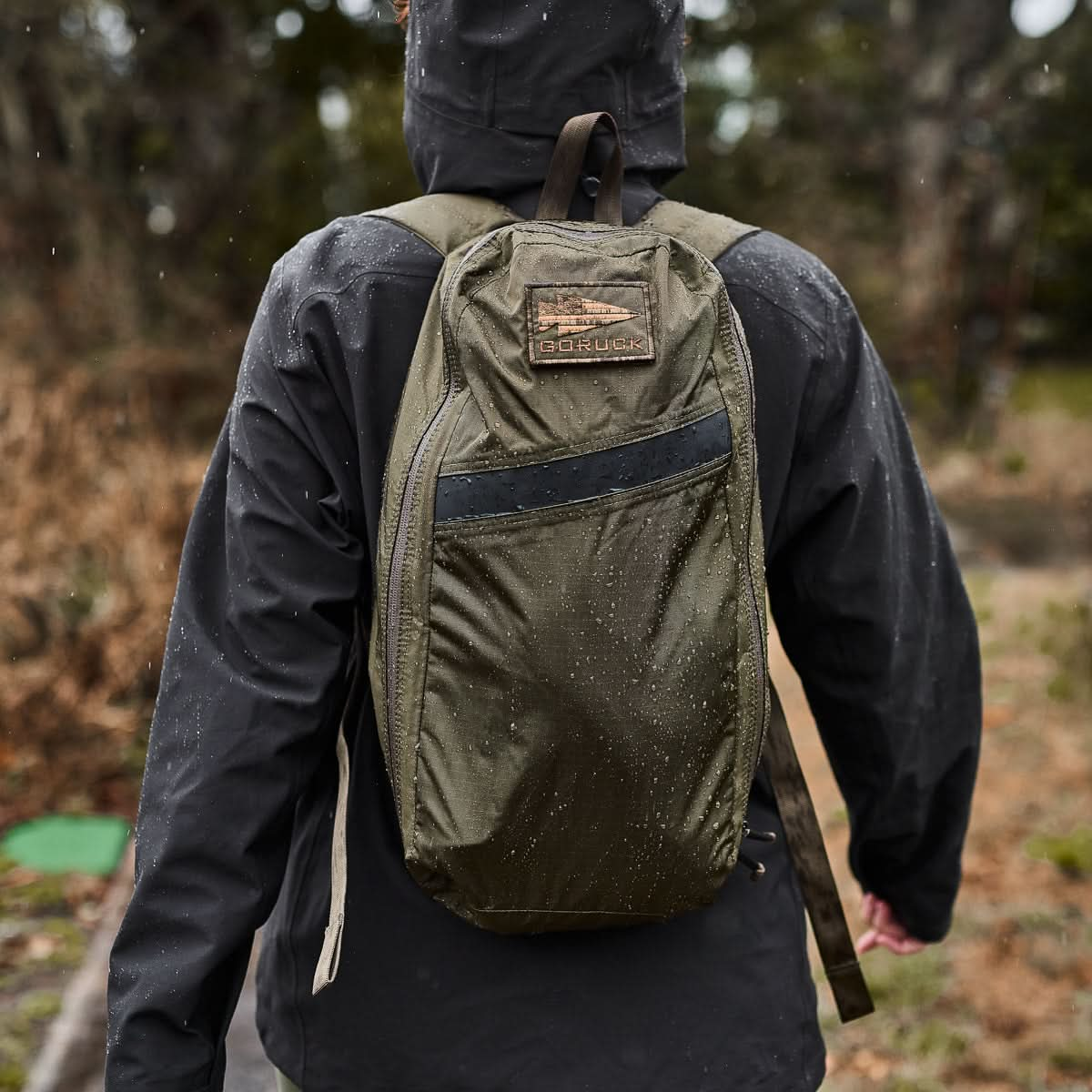 A person in a black jacket strolls along a trail, their Packable Bullet Ruck - Ripstop ROBIC® backpack glistening with light rain.