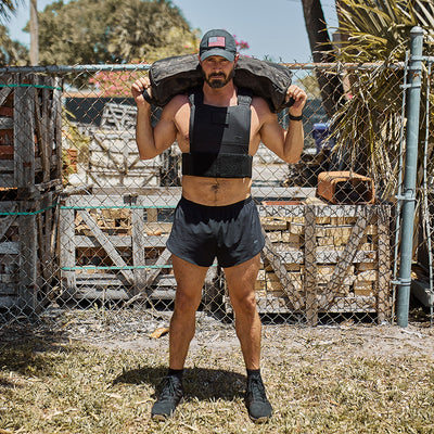 A man stands outdoors, demonstrating his strength with a large sandbag draped over his shoulders. He wears a cap, durable black vest, and GORUCK Men’s Ranger Panties made from ToughStretch fabric. Positioned on dry grass near a chain-link fence and trees, his outfit epitomizes durability similar to the Scars Lifetime Guarantee.