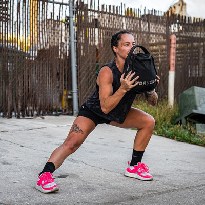 Holding a black weighted bag, a person performs a lateral lunge outdoors. They are sporting GORUCK's Women's Rough Runner - Hot Pink lightweight running shoes, paired with a black tank top and shorts. The vibrant pink of their shoes stands out against the fence and surrounding greenery.