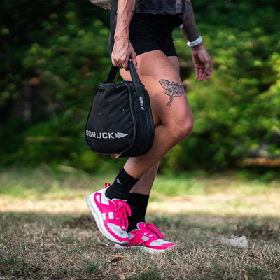 A person sporting GORUCK's Women's Rough Runner in Hot Pink lightweight running shoes and black shorts carries a black GORUCK bag. A butterfly tattoo adorns their leg as they stand outdoors, surrounded by trees, making it an ideal setting for a Rough Runner™ challenge.