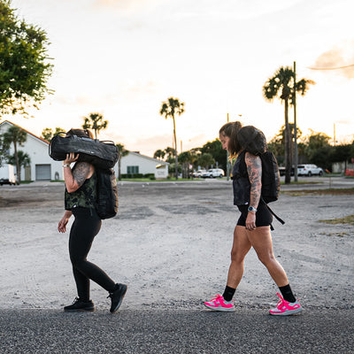 Two people stroll along a rural road at sunset, carrying large backpacks. Wearing Women's Rough Runner shoes in Hot Pink by GORUCK and sporting athletic gear like leggings and shorts, they move gracefully past palm trees and buildings in the background.