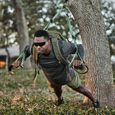 In a park-like setting, a man dressed in workout gear and sunglasses is using the GORUCK X TRX BUNDLE attached to a tree for an incline push-up exercise. His concentration turns the greenery and trees around him into his personal home gym.