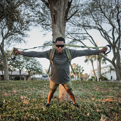 A person works out outdoors with the GORUCK X TRX BUNDLE from GORUCK, attaching the suspension trainer to a tree. They stretch their arms wide while pulling on the bands, surrounded by grass, trees, and houses under a clear sky.