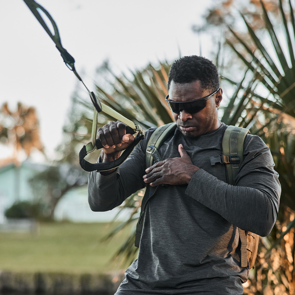 A person sporting sunglasses and a dark long-sleeve shirt works out outdoors with the GORUCK X TRX BUNDLE from GORUCK. Their expression is determined as they exercise amid palm trees, with their home gym in the background.