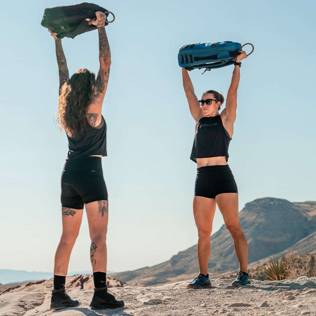 Two individuals exercising outdoors, hoisting GORUCK Rucker 4.0 bags overhead against a mountainous backdrop.