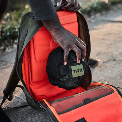 A person places a black mesh Suspension Trainer pouch from the GORUCK X TRX BUNDLE into a brightly colored orange GORUCK backpack on a stone surface. The open backpack reveals organized compartments ideal for rucking essentials, while sunlight casts a warm glow on the scene.