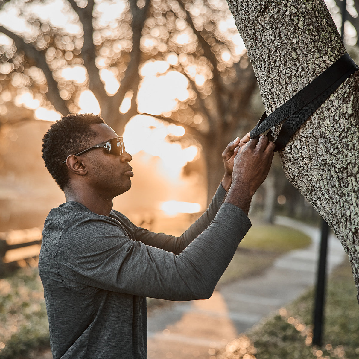 A person wearing sunglasses uses the GORUCK X TRX BUNDLE to secure a suspension trainer strap around a tree trunk in the park. As the sun sets, it casts a warm glow over the scene. In the background, a path and benches offer an ideal setting for those enjoying their outdoor home gym routine with GORUCK equipment.