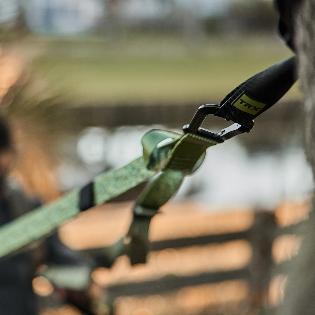 A close-up of a GORUCK X TRX BUNDLE secured to a tree outdoors shows the strap with a metal clip and green fabric band, perfect for home gym enthusiasts looking for versatile fitness solutions. In the background, there's a blurred figure and wooden fence set against a natural backdrop.