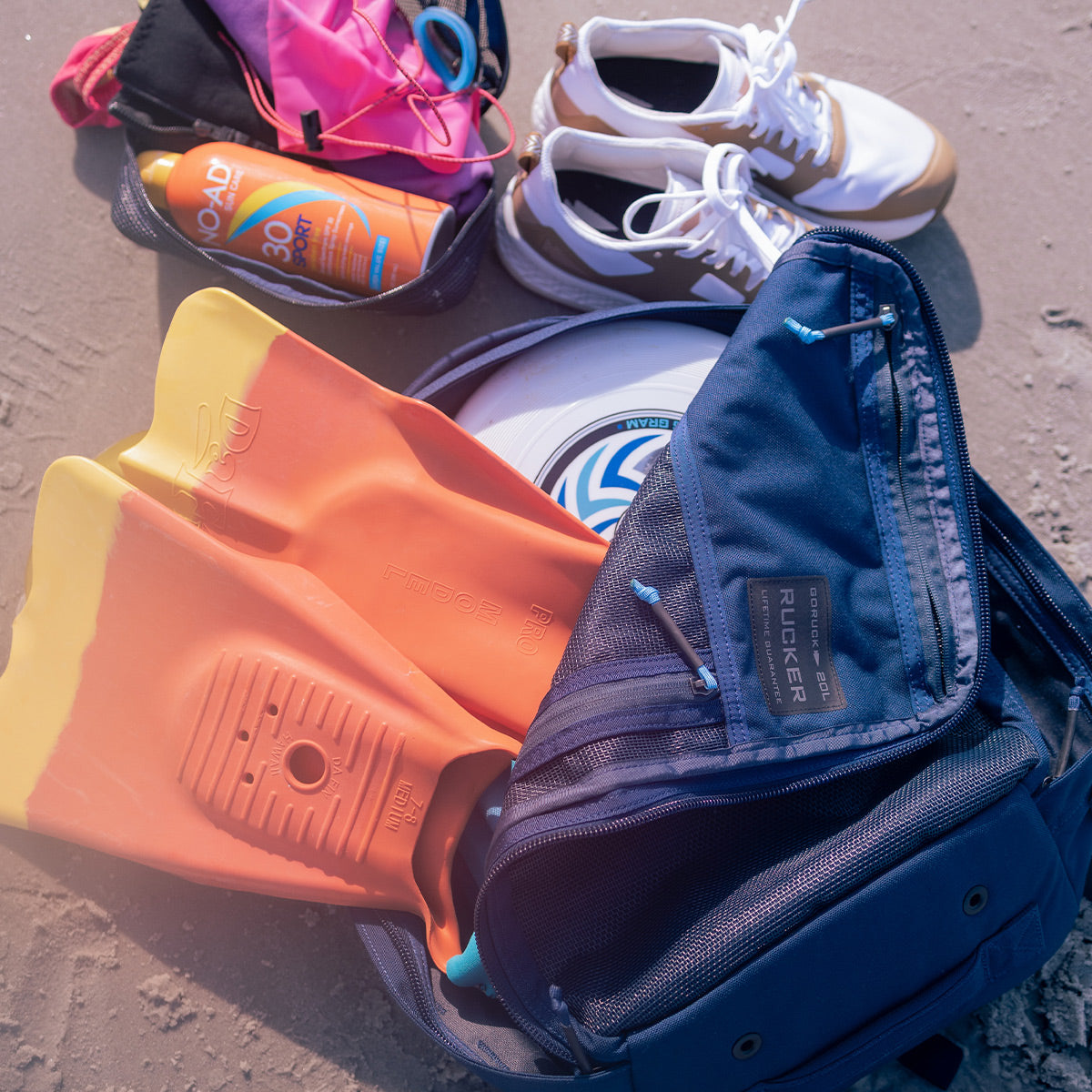 On the sandy beach lies a Rucker 4.0 20L in Navy with its distinctive Navy Mesh Front Slant, surrounded by orange flippers, a frisbee, sunscreen, and white sneakers. A pink bag peeks from the pack as gentle sunlight creates soft shadows, inspiring thoughts of future rucking adventures with GORUCK gear.