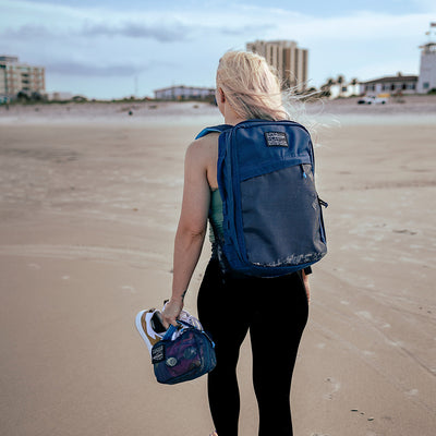 A person with long hair, participating in rucking, strolls along a beach carrying a substantial Rucker 4.0 20L - Navy + Navy Mesh Front Slant backpack from GORUCK on their back and a smaller vibrant bag in one hand. In the distance, buildings rise against the cloudy sky backdrop.