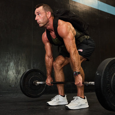 In a dimly lit gym, a man in white sneakers and a smartwatch performs a deadlift while wearing the Rucker 4.0 weighted vest by GORUCK. With the focus of GORUCK enthusiasts, he embodies the spirit of rucking as he lifts the barbell, standing against a wall marked by its distinctive blue stripe.