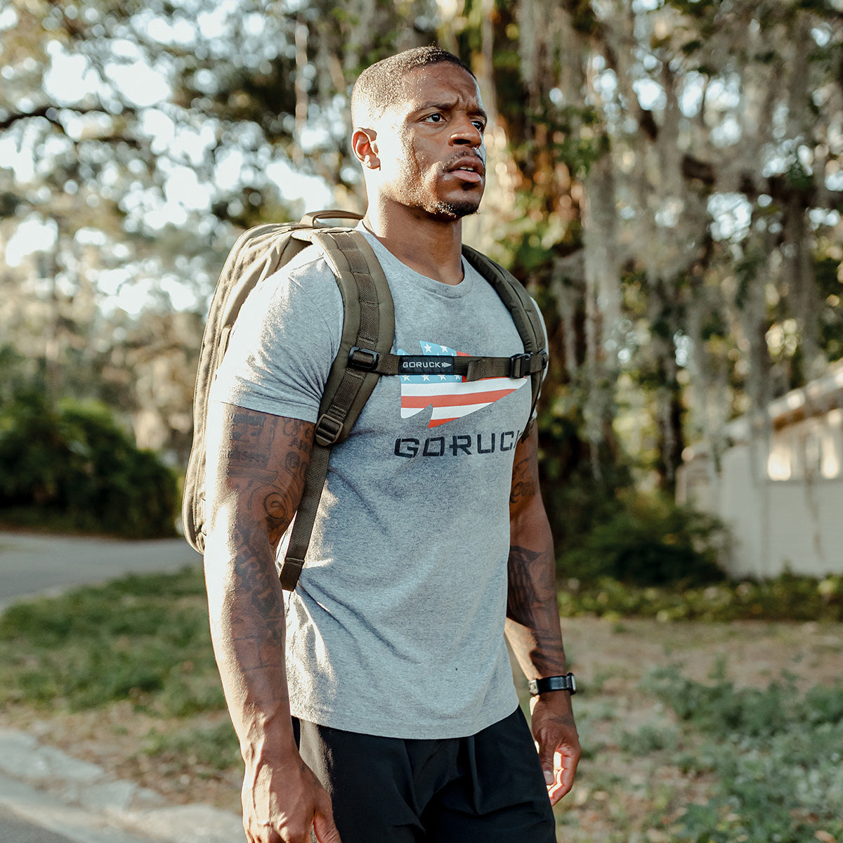 A man strolls outdoors with a sizable GORUCK Rucker 4.0 backpack. Clad in a gray t-shirt with a logo, black shorts, and a smartwatch, he embodies the spirit of rucking. The backdrop showcases trees and a blurred residential area, hinting at a suburban setting.