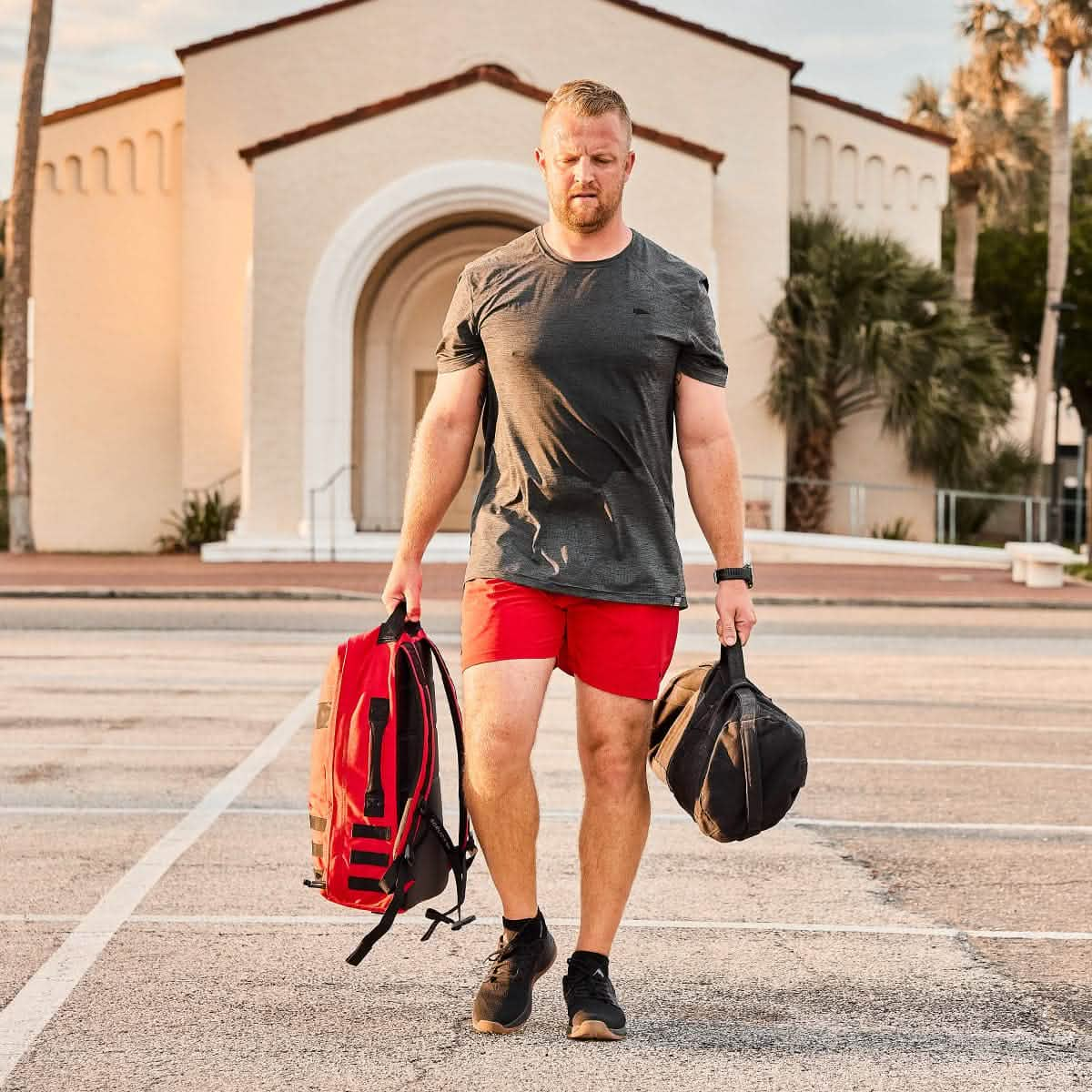 A man in red shorts, carrying bags weighted like GORUCK Rucker 4.0 plates, strides through an empty parking lot. The white building looms in the background as he embodies the spirit of rucking, turning an ordinary day into a fitness journey.