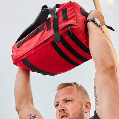 A person lifts a GORUCK Rucker 4.0 overhead, tattoos adorning their arms and a wristwatch on their wrist, embodying the spirit of rucking with every powerful movement.