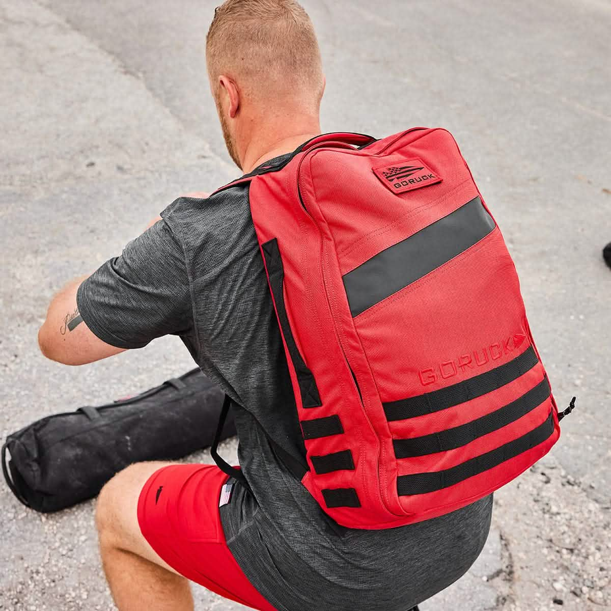 A person in a gray shirt and red shorts is wearing a GORUCK Rucker 4.0 backpack with ruck plates while crouching on a paved surface.