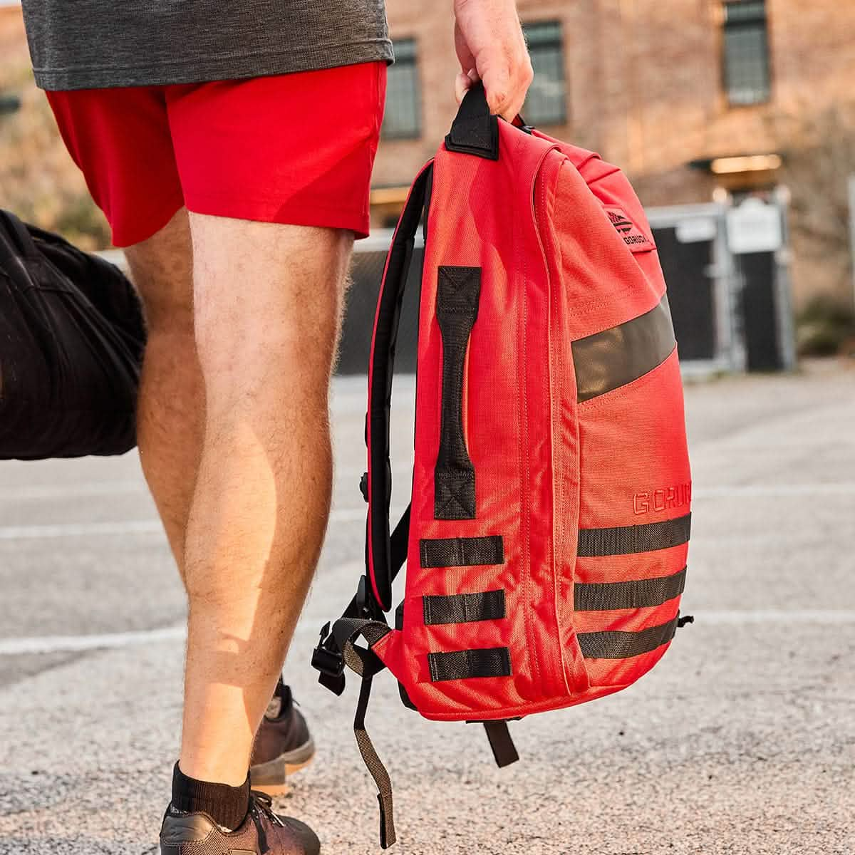 A person with a GORUCK Rucker 4.0 red backpack, wearing red shorts and black sneakers, walks outside, ready for a rucking adventure.