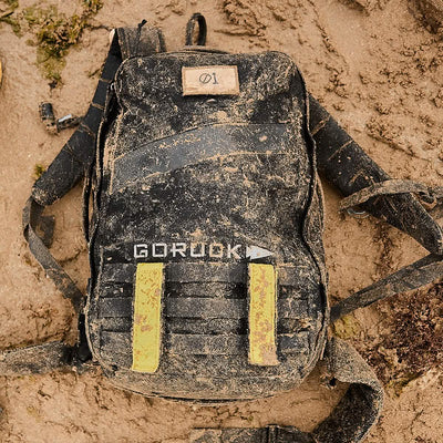 A GORUCK Rucker 4.0 in a muddy black color with yellow straps rests on the sandy ground, making it perfect for any rucking adventure.