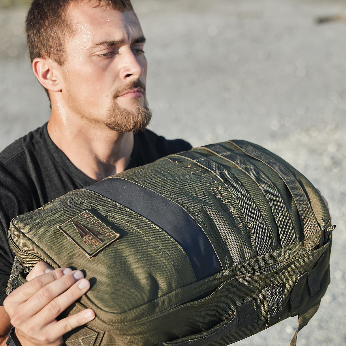 A person in a black shirt, sweating, holds a sturdy GORUCK Rucker 4.0 with black accents. The setting appears to be outdoors, with sunlight highlighting the intensity of their rucking adventure.