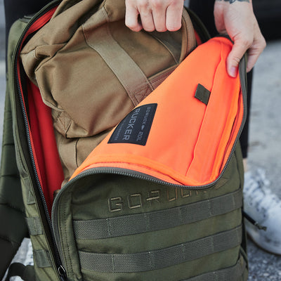 A person opens an olive green Rucker 4.0 by GORUCK, revealing its bright orange interior. The bag features a brown internal compartment and distinct label details. One hand grips the zipper, and part of a shoe is visible in the background, suggesting an active day of rucking ahead.