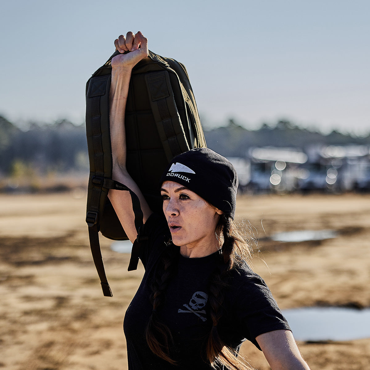 A woman wearing a black beanie and t-shirt proudly holds the GORUCK Rucker 4.0 aloft with one hand, standing outdoors on a sandy area with puddles and trees in the background.