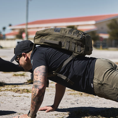 A man in a black cap and T-shirt performs a push-up outdoors on a sandy surface, rucking with the Rucker 4.0 backpack by GORUCK in olive green. Tattoos adorn his arm as he focuses on his workout, while a building with a red roof stands in the background.