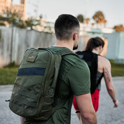 Two individuals are rucking along a street, each equipped with a Rucker®. The person at the front is wearing a green shirt paired with an olive GORUCK Rucker 4.0, while the one behind has on red shorts and carries a black backpack. A wooden fence and trees form the backdrop of their route.