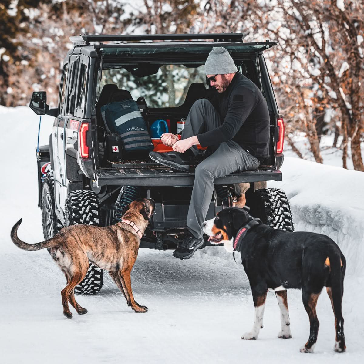 A man sits on the tailgate of an SUV in the snow, pulling on boots with two dogs at his side—ready for a rucking adventure with his reliable Rucker 4.0.