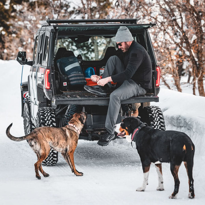 A man sits on the tailgate of an SUV in the snow, pulling on boots with two dogs at his side—ready for a rucking adventure with his reliable Rucker 4.0.