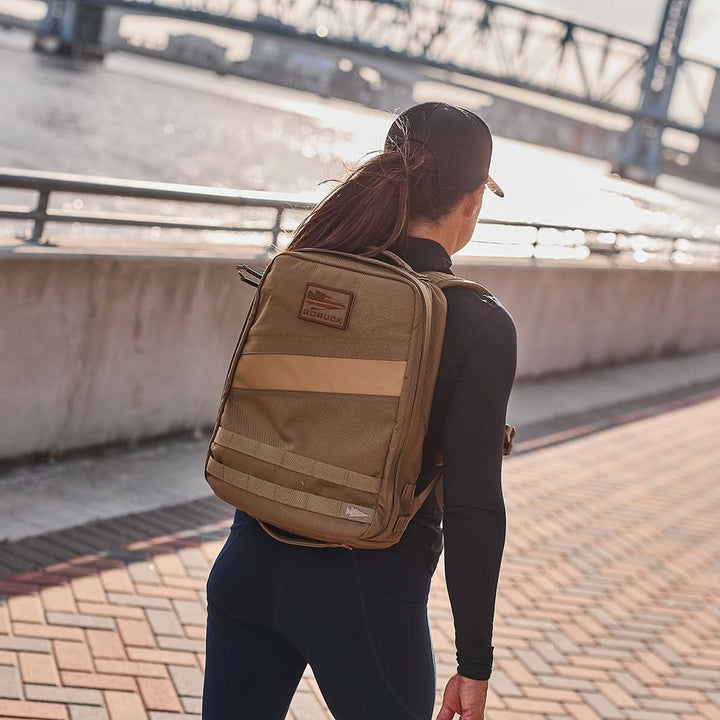 A woman in athletic wear goes rucking by a river with a tan Rucker 4.0 backpack, passing near a bridge and railing.