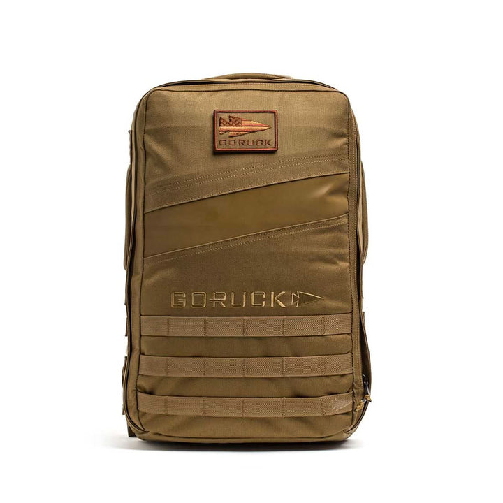 A tan Rucker 4.0 rucksack with a front patch, horizontal straps, and zipper compartments stands upright—perfect for rucking adventures.