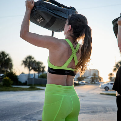 During sunset, a woman dressed in the GORUCK Power Bra, featuring Toughflex technology, lifts a weighted bag outdoors. Palm trees and buildings provide a stunning backdrop.