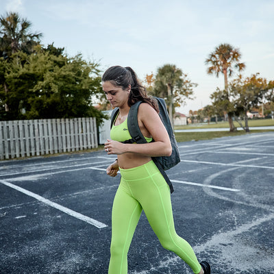 A woman in vibrant green athletic wear featuring the durable Toughflex fabric from GORUCK's Power Bra collection sprints through a parking lot, with trees and a fence providing the backdrop under an overcast sky.