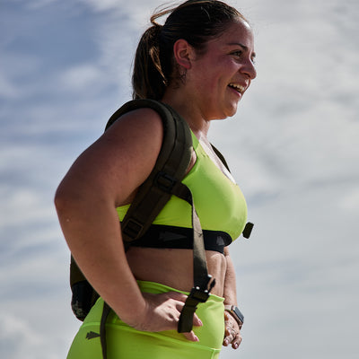 A woman wearing vibrant green Power Bra - Toughflex, crafted with GORUCK's signature ToughStretch Fabric for high-impact movements, smiles while standing outdoors. She has a backpack and places her hands on her hips. The sky is visible in the background.