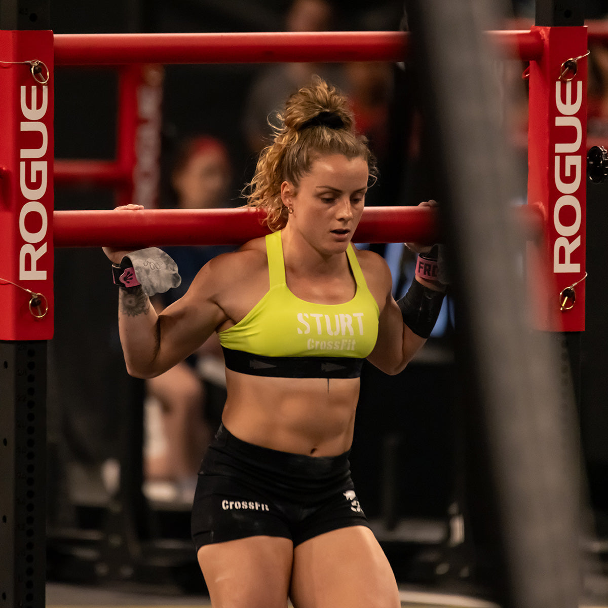 A woman performs a squat exercise with a heavy barbell on her shoulders, highlighting the Toughflex Fabric of her bright yellow Power Bra by GORUCK. At the gym, under the Rogue branding, she stays focused and prepared for more high-impact movements, with her hair tied up and sporting black shorts.