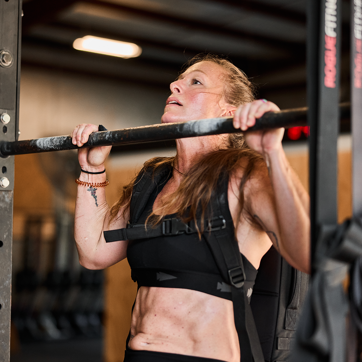 A woman in athletic gear is doing a pull-up in a gym, wearing the GORUCK Power Bra - Toughflex and a weighted vest. Her determined expression reflects her focus on high-impact movements, surrounded by various exercise equipment.