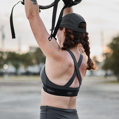 A woman with braided hair and a baseball cap is exercising outdoors, engaging in high-impact movements with suspension straps held above her head. She is wearing the GORUCK Power Bra - Toughflex along with leggings made from ToughStretch Fabric, set against a backdrop of blurred trees and a cloudy sky.