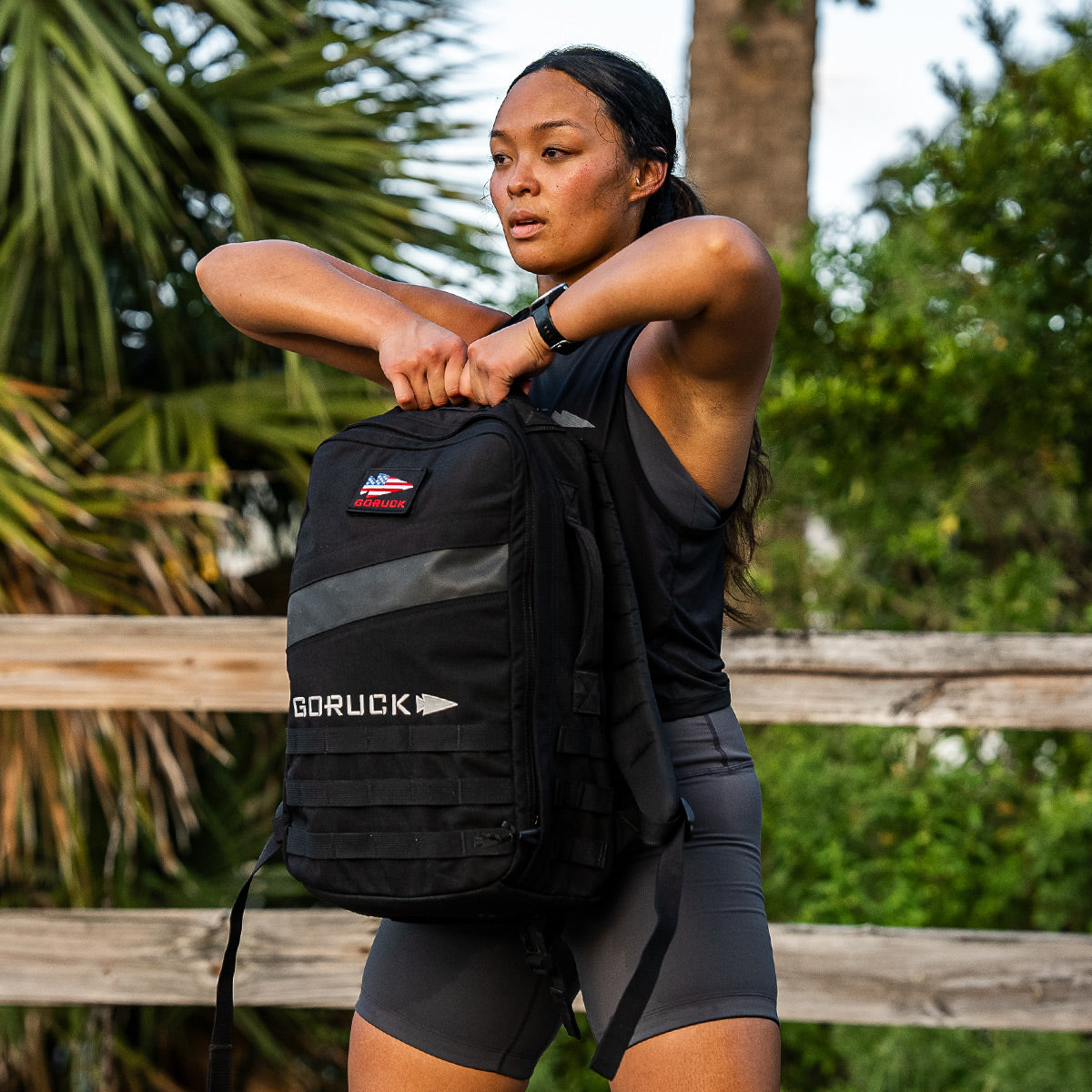 Standing outside near a wooden fence, a person adjusts a black GORUCK backpack. Dressed in the GORUCK Power Bra - Toughflex and shorts designed for high-impact movements, they exude readiness amidst the lush green foliage in the background.