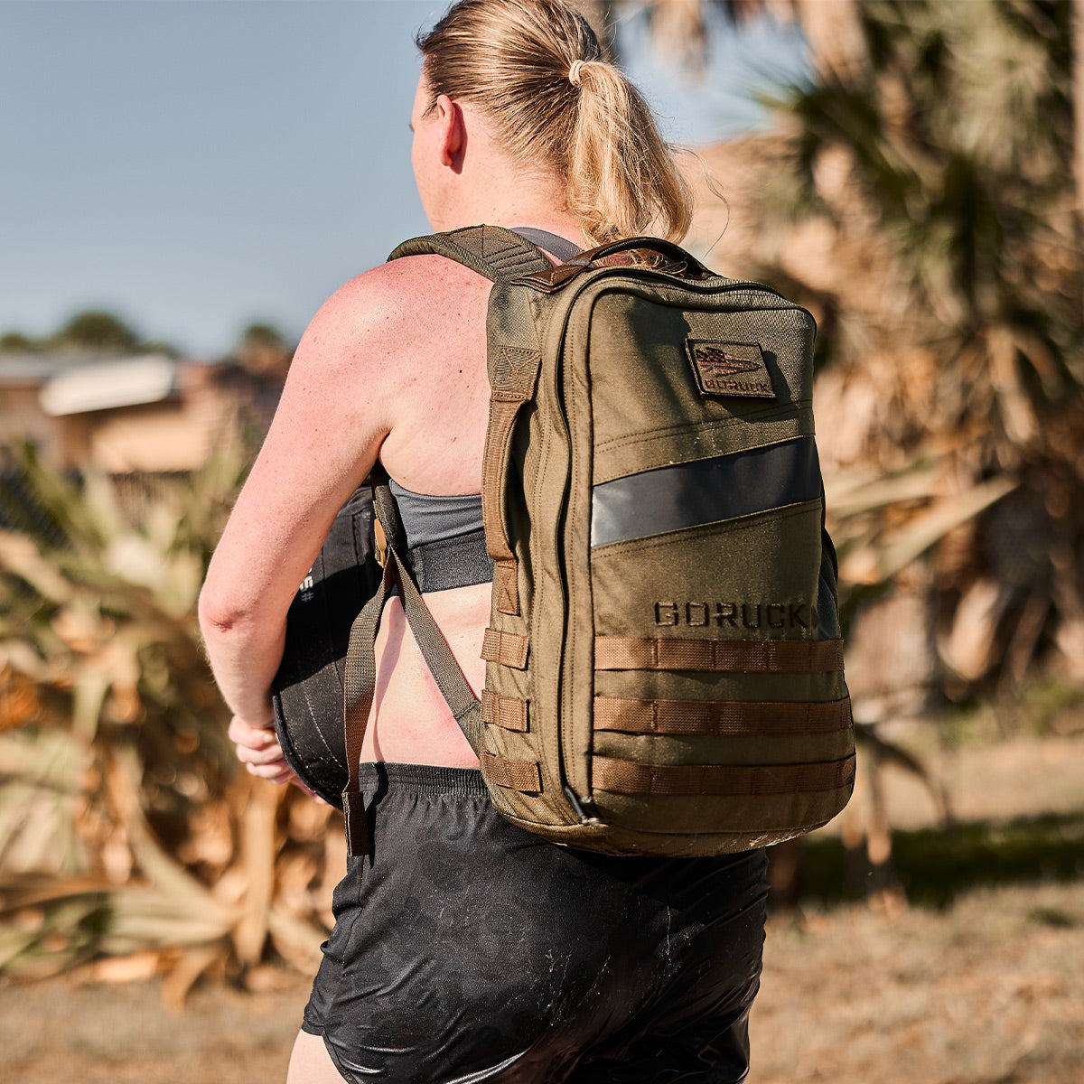 A person wearing a green tactical backpack is engaged in an outdoor activity, dressed in a gray tank top and black shorts designed for high-impact movements. The GORUCK Power Bra - Toughflex offers support as it peeks from underneath their top while they move confidently amidst the trees and the nearby building.