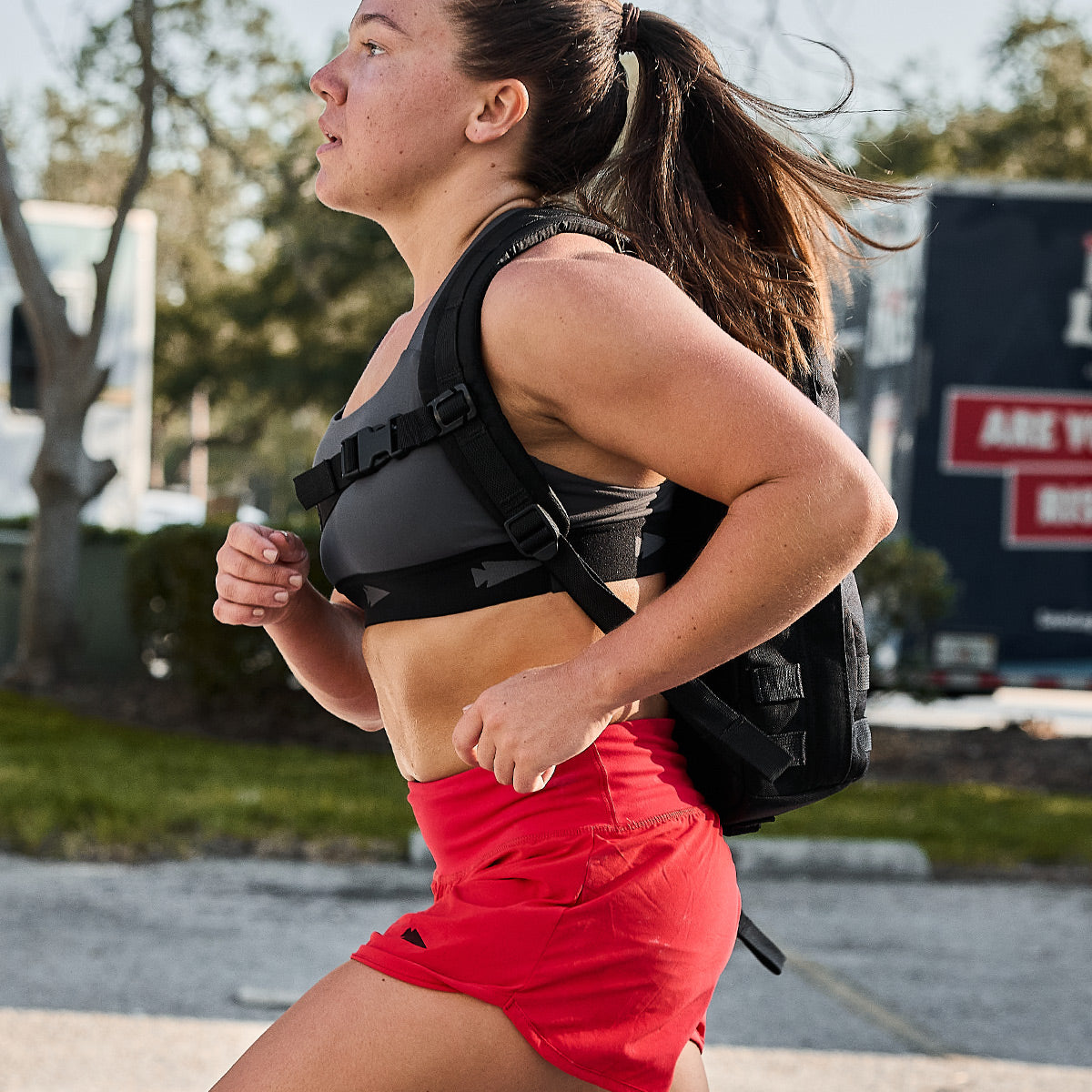 A woman in a GORUCK Power Bra - Toughflex and red shorts is running outdoors, her black backpack securely in place. She appears focused, with her hair tied back, navigating the trees and a building in the background under a clear sky.