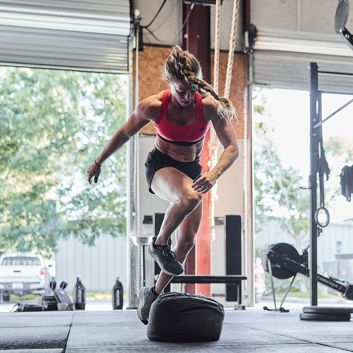 A woman wearing athletic attire, including the GORUCK Power Bra - Toughflex, is captured mid-jump over a large tire in a gym with open garage doors. Sunlight pours in, accentuating her high-impact movement amidst CrossFit equipment visible in the background.