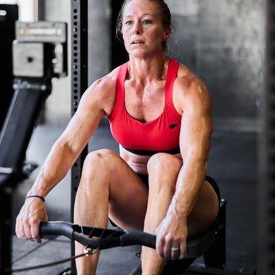 A woman in a GORUCK Power Bra - Toughflex and black shorts is intensely rowing in the gym. Her sweat-drenched focus highlights her determination during these high-impact movements, with a blurred background emphasizing her posture and grit.