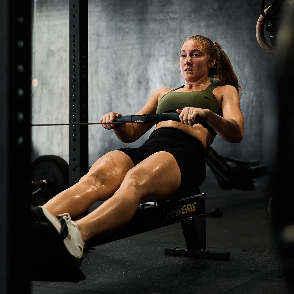 A woman dressed in athletic wear, featuring the Power Bra made from Toughflex by GORUCK, intensely uses a rowing machine at a gym. Her hands firmly grip the handle as her expression reveals focus and determination amid weights and exercise equipment crafted for high-impact workouts.