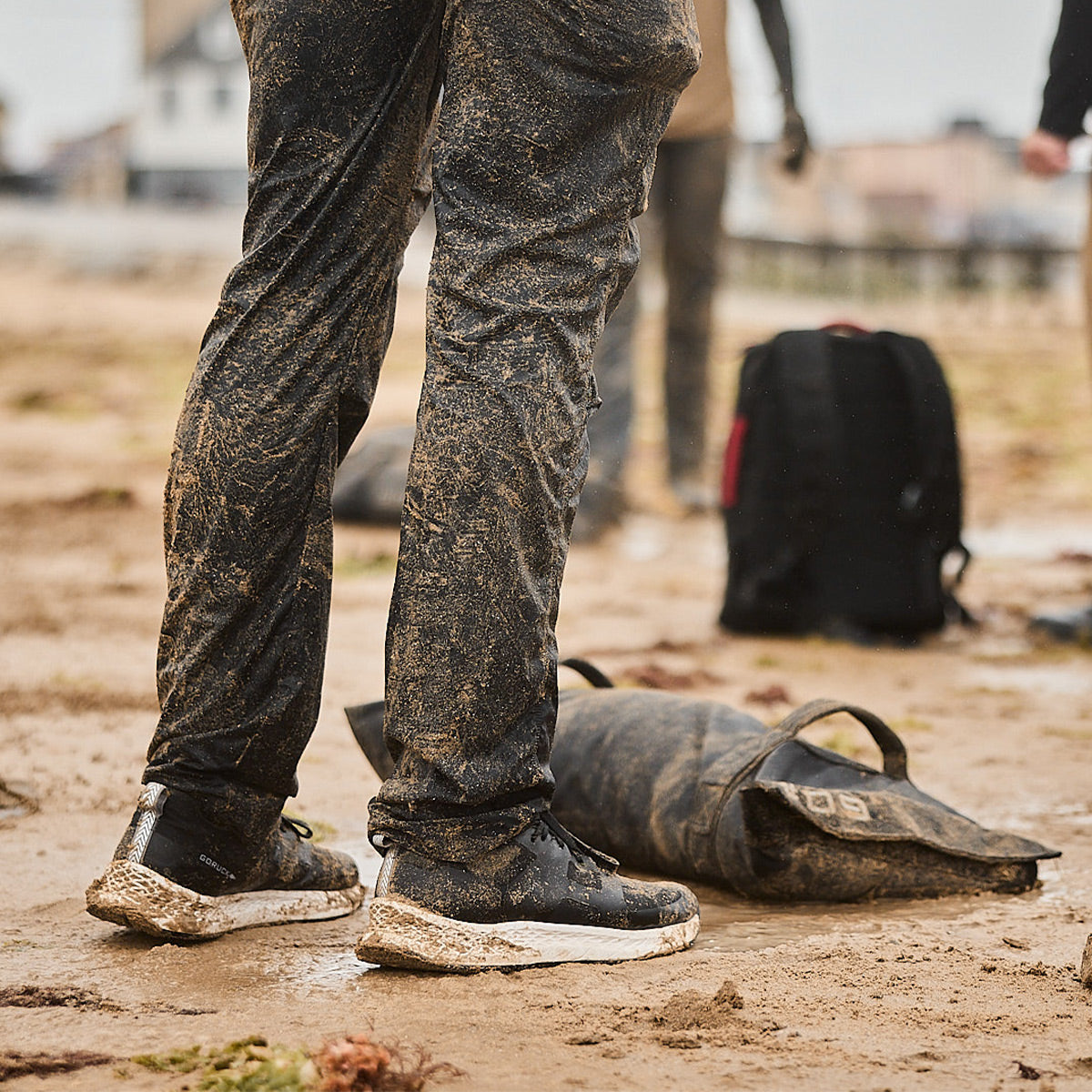 A person stands on a muddy surface, their dirty pants and shoes hinting at the adventures that come with wearing the Men's Rough Runner - Black + White by GORUCK. Nearby lies a large, mud-caked bag. In the blurred background, another figure paired with a black GORUCK backpack maintains an air of Special Forces mystique.