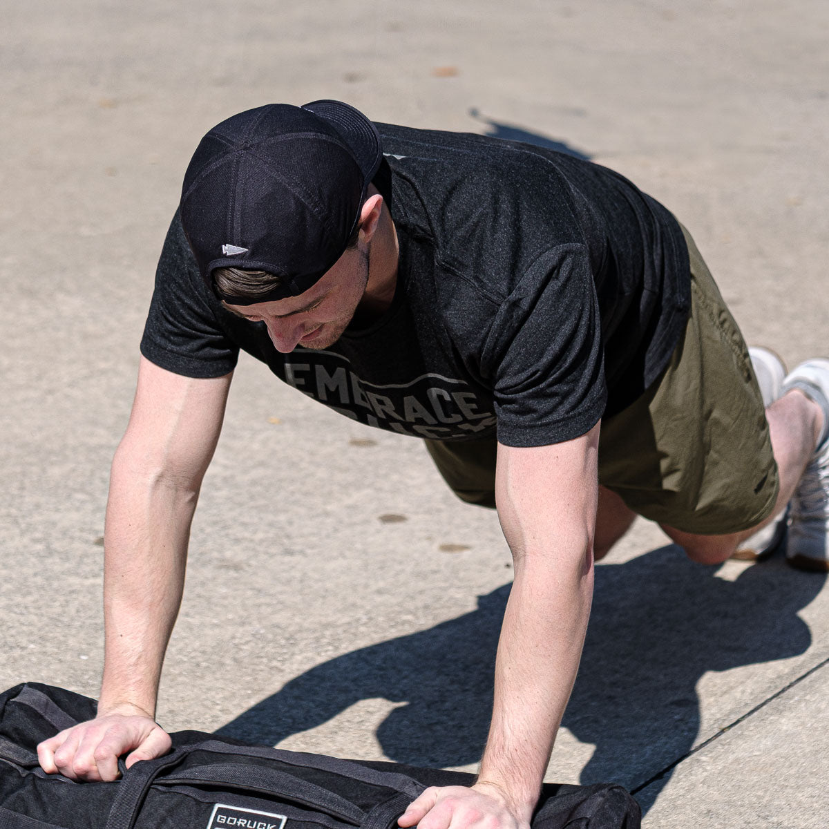 Outdoors, a person does push-ups on a sandbag, sporting a black Performance Running Hat made of ToughMesh fabric, along with a black shirt and green shorts.