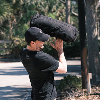 Amidst lush greenery, a man in a Performance Running Hat - ToughMesh is lifting a heavy sandbag outdoors, seamlessly combining strength with style.