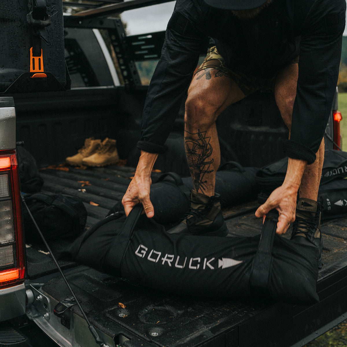 A person lifts a Simple Training Sandbag by GORUCK onto the bed of a pickup truck, amidst scattered gear and boots. Dressed in a black jacket and camo shorts, with tattoos visible on their leg, they depend on the durability ensured by the Scars Lifetime Guarantee.