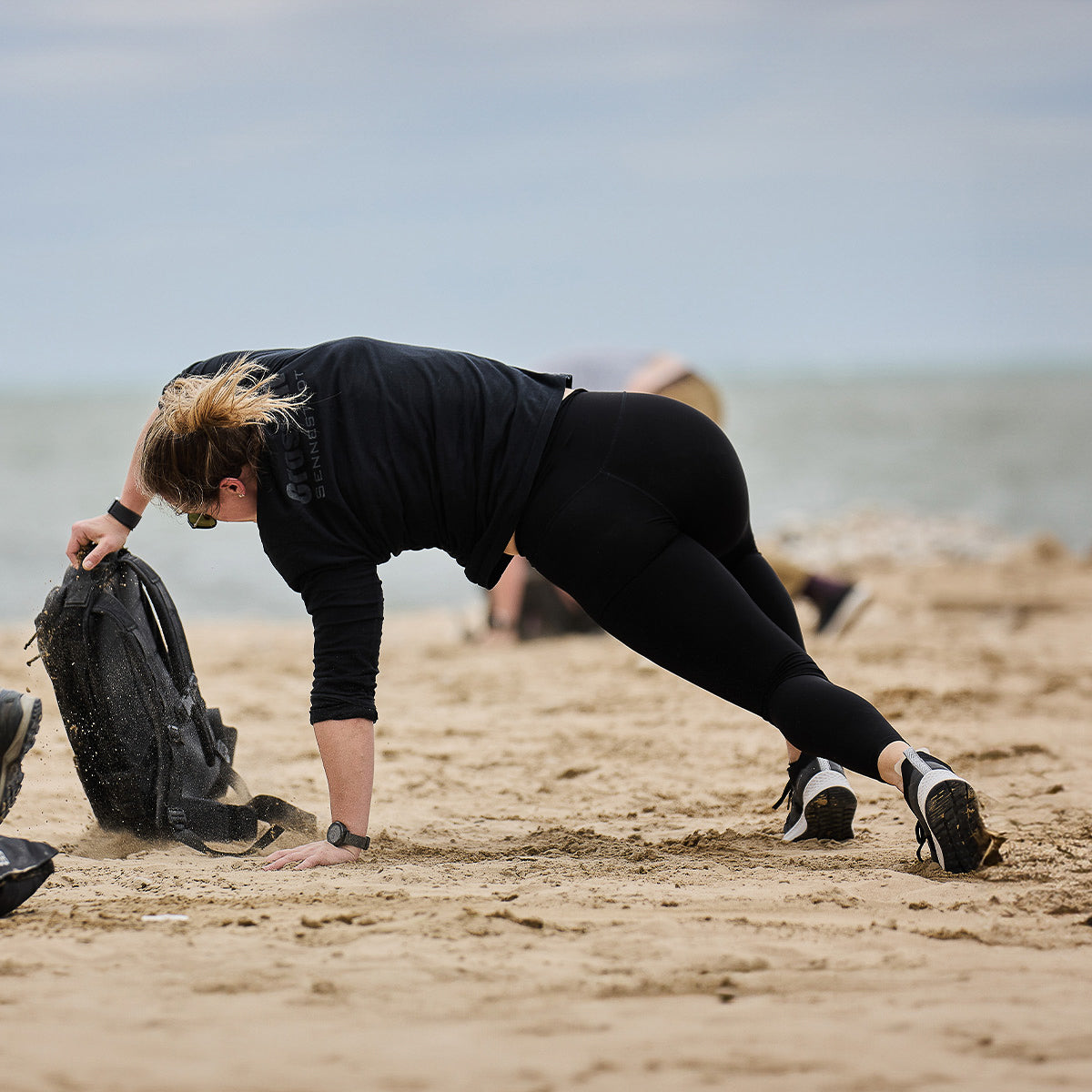 Dressed in athletic gear, a person skillfully performs a side plank on the sandy beach while lifting a weighted bag labeled "Women's Rough Runner - Black + White" by GORUCK. The ocean extends behind them under a sky adorned with thick clouds.