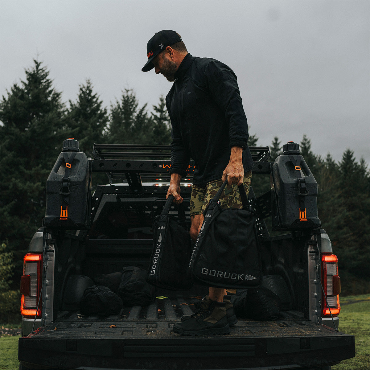 A man in a black cap and camo shorts stands in the bed of a pickup truck, gripping his GORUCK Sand Jerry Can crafted from Special Forces Grade materials. Forested trees form the backdrop on this cloudy day, hinting at the rugged journey ahead.